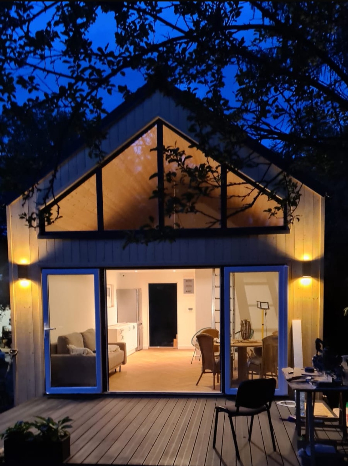 Oświetlony dom szkieletowy z dużym oknem trójkątnym i otwartymi drzwiami balkonowymi, widok z zewnątrz na drewniany taras i wnętrze z meblami.
