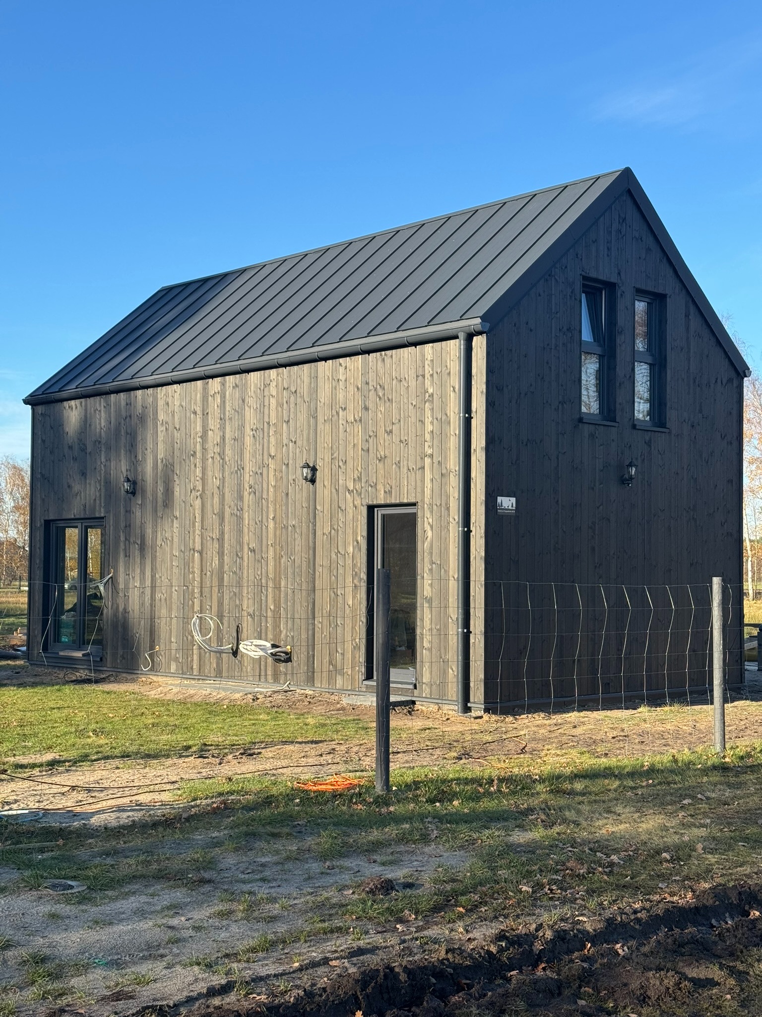 Nowoczesny, dwukondygnacyjny dom z ciemnego drewna, z metalowym dachem w kolorze grafitu, oświetleniem zewnętrznym i ogrodzeniem z siatki, na tle błękitnego nieba.