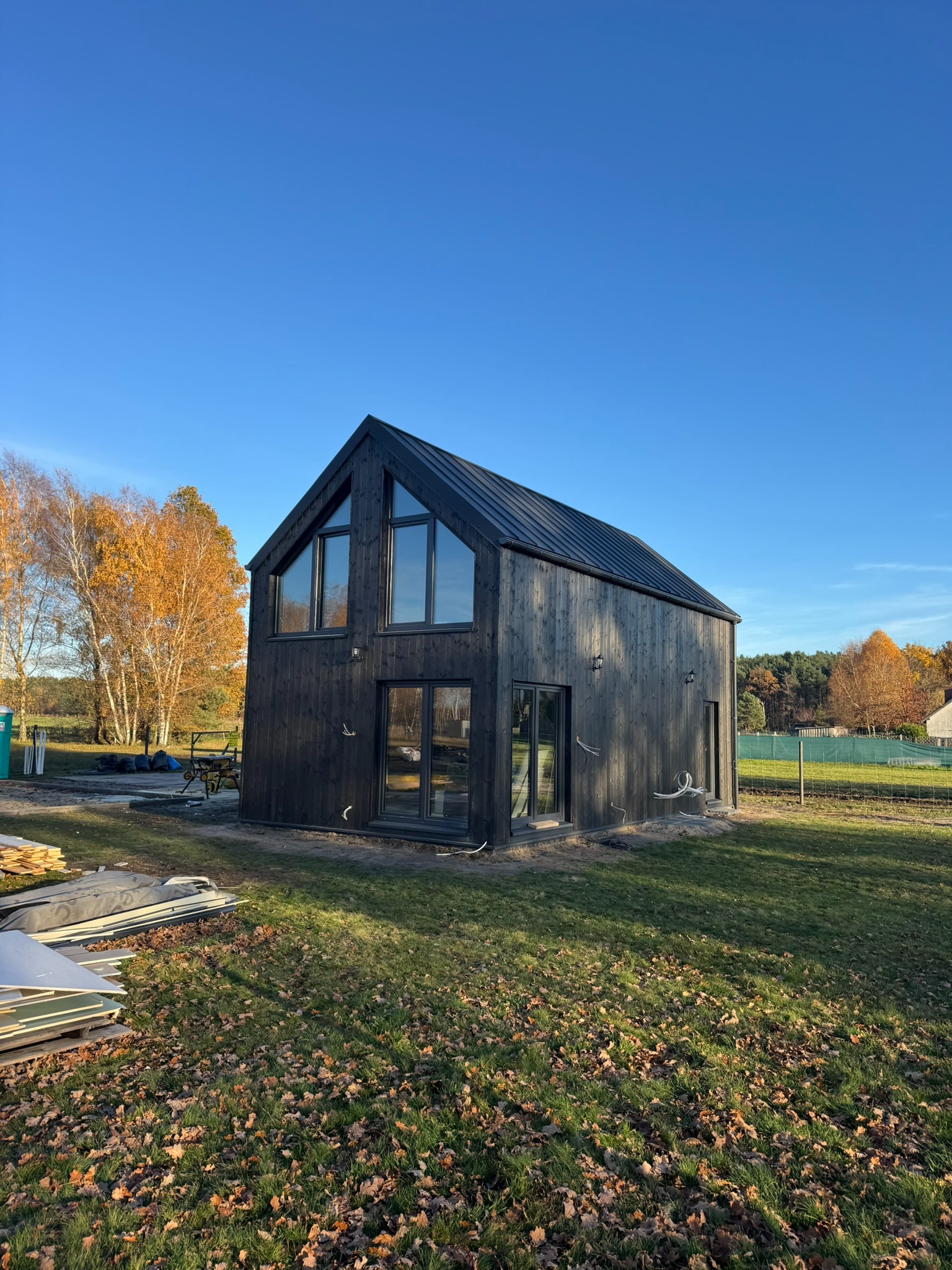 Czarny, nowoczesny dom szkieletowy z dużymi oknami, otoczony zielonym trawnikiem i jesiennymi drzewami pod błękitnym niebem. Widoczne materiały budowlane obok.