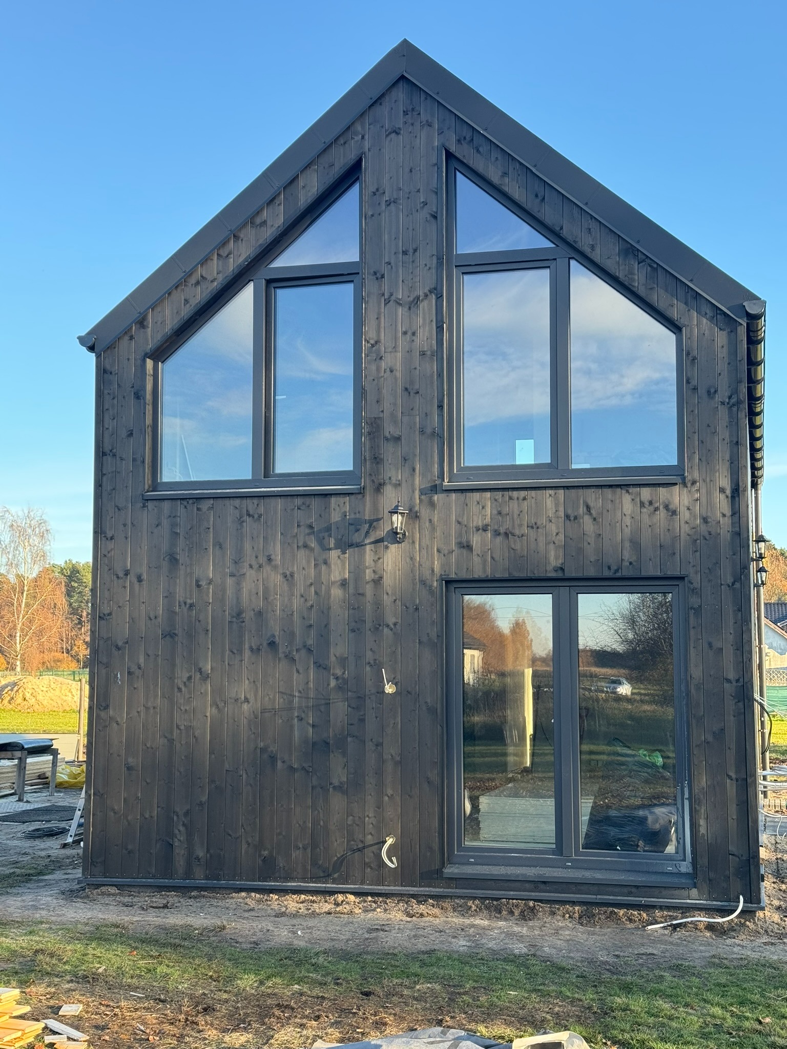 Dwukondygnacyjny dom szkieletowy z ciemnego drewna, z dużymi oknami w kształcie trójkątów na górze i prostokątnymi na dole, odbijającymi błękit nieba.