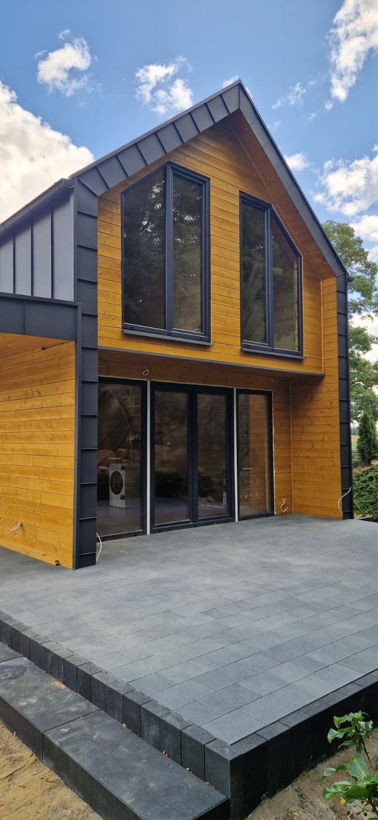 Nowoczesny dom z drewna z dużymi oknami i tarasem wyłożonym szarą kostką. Elewacja w odcieniach brązu i czerni. Widok na błękitne niebo z chmurami.