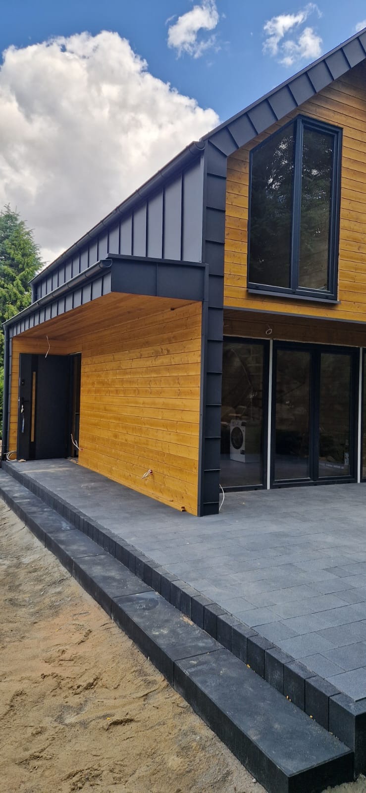 Nowoczesny dom z drewna z szarym dachem i czarnymi ramami okiennymi. Widoczny taras z kostki brukowej i fragment ogrodu. Jasne drewno elewacji.