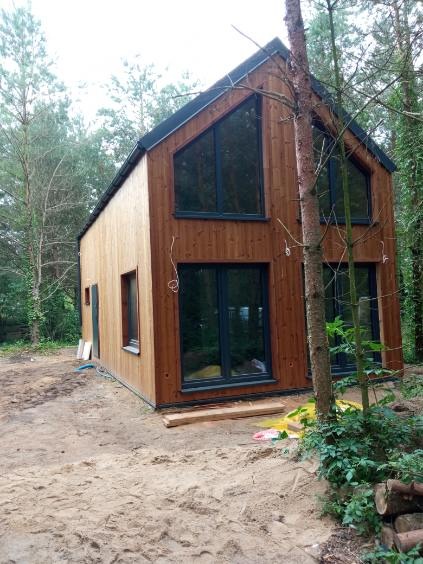 Nowoczesny, dwukondygnacyjny dom z drewna, z dużymi oknami i czarną stolarką, otoczony drzewami. Widoczne elementy wykończeniowe i przygotowany teren.