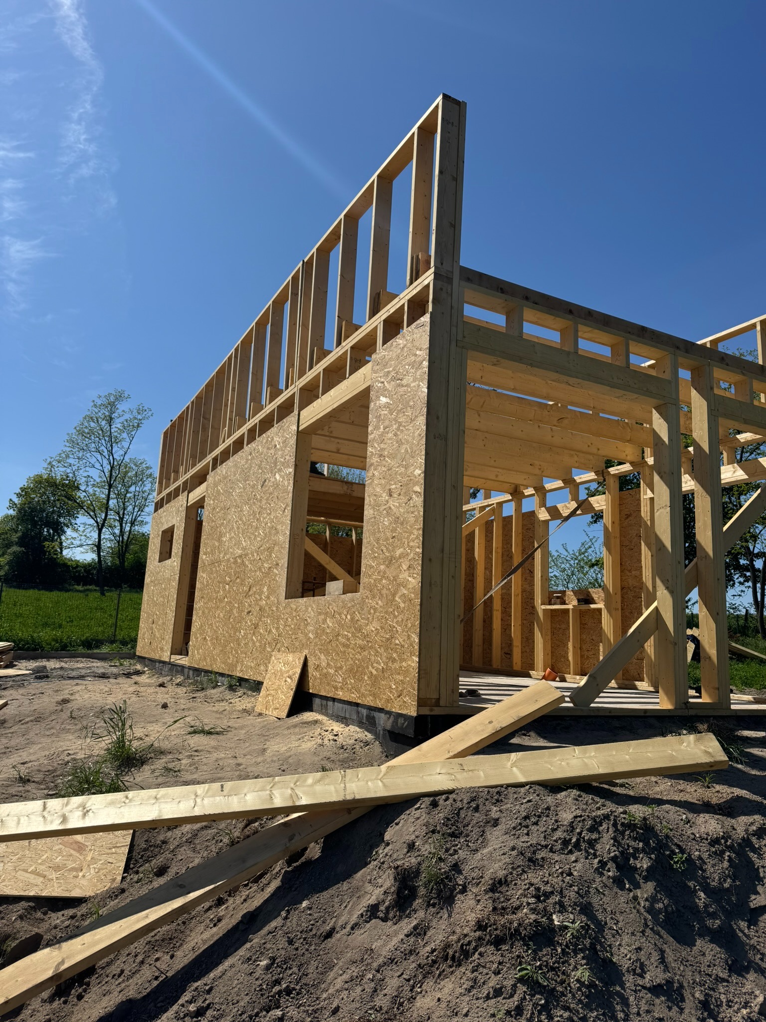 Konstrukcja szkieletowa domu w trakcie budowy, widok z dołu na drewniane elementy i płyty OSB, błękitne niebo w tle. Widoczne deski na ziemi.