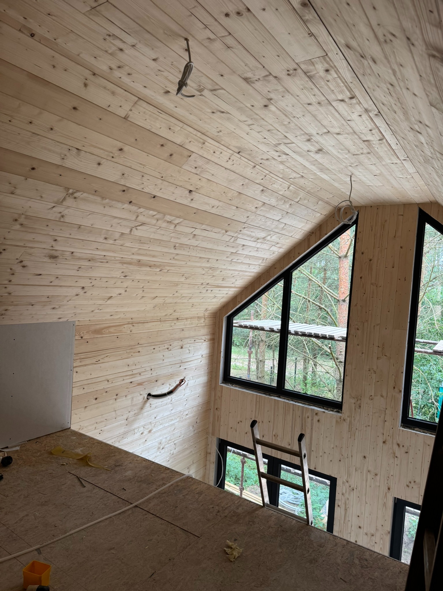 Wnętrze domu szkieletowego w trakcie wykańczania: drewniane ściany i sufit, okna z widokiem na zewnątrz, drabina. Widoczne elementy instalacji elektrycznej.
