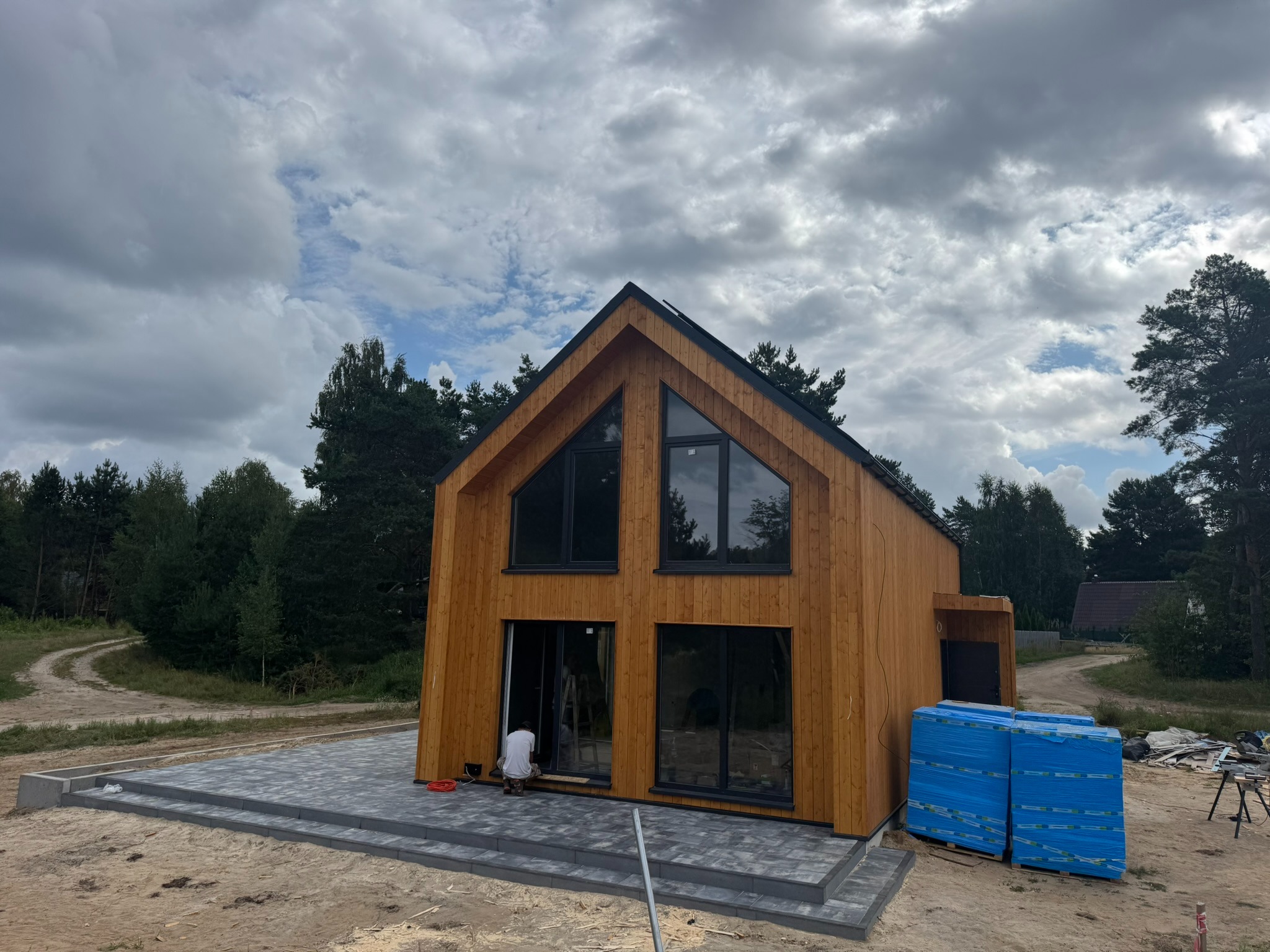 Nowoczesny dom szkieletowy z drewnianą elewacją i dużymi oknami, częściowo wykończony taras z kostki brukowej, widoczne materiały budowlane i pracownik przy drzwiach.