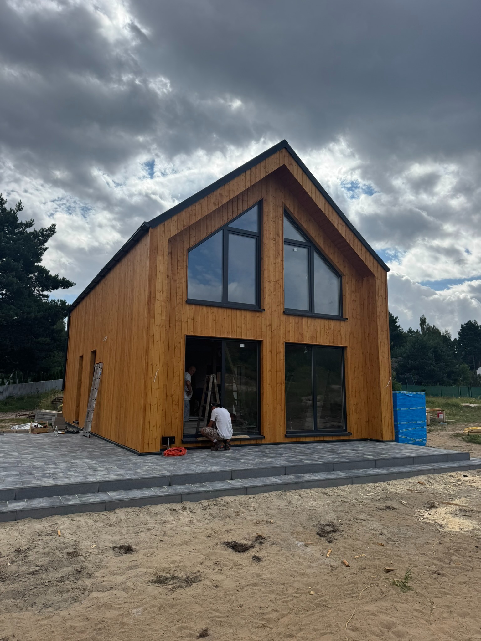 Nowoczesny dom z drewna w trakcie wykończenia, z dużymi oknami i tarasem z kostki. Widoczni pracownicy wewnątrz, pochmurne niebo w tle.