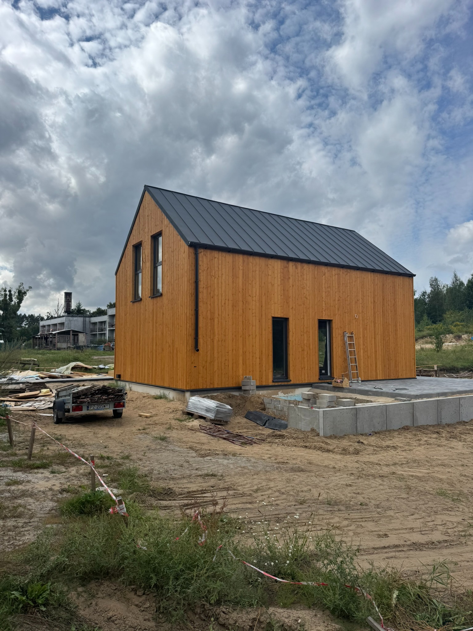 Nowoczesny, drewniany dom z ciemnym dachem, częściowo wykończony, z widocznymi fundamentami i przyczepką z materiałami budowlanymi w tle.