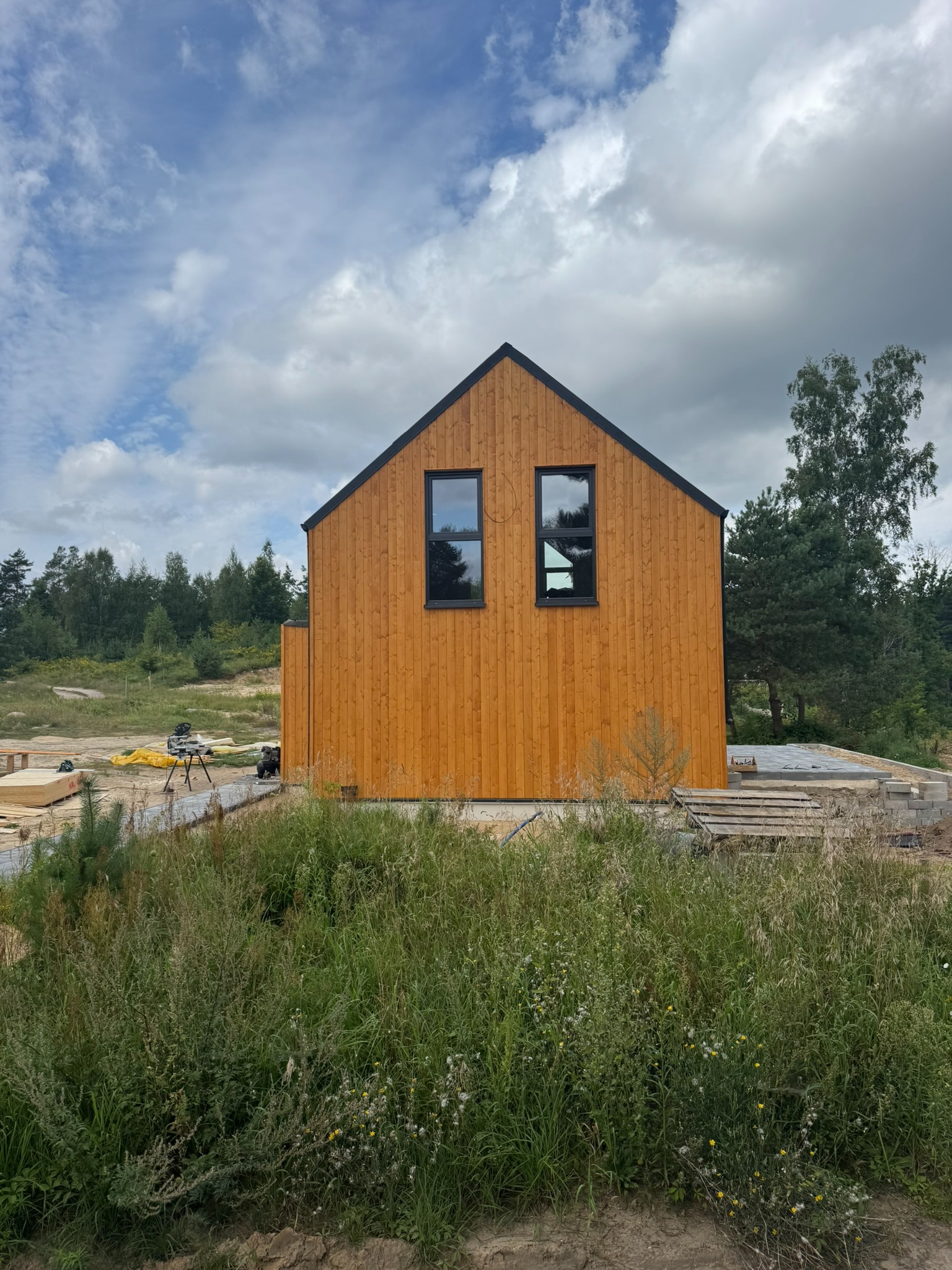 Surowy, drewniany dom szkieletowy z czarnymi ramami okiennymi, w otoczeniu zieleni i placu budowy. Widoczne narzędzia i materiały budowlane.