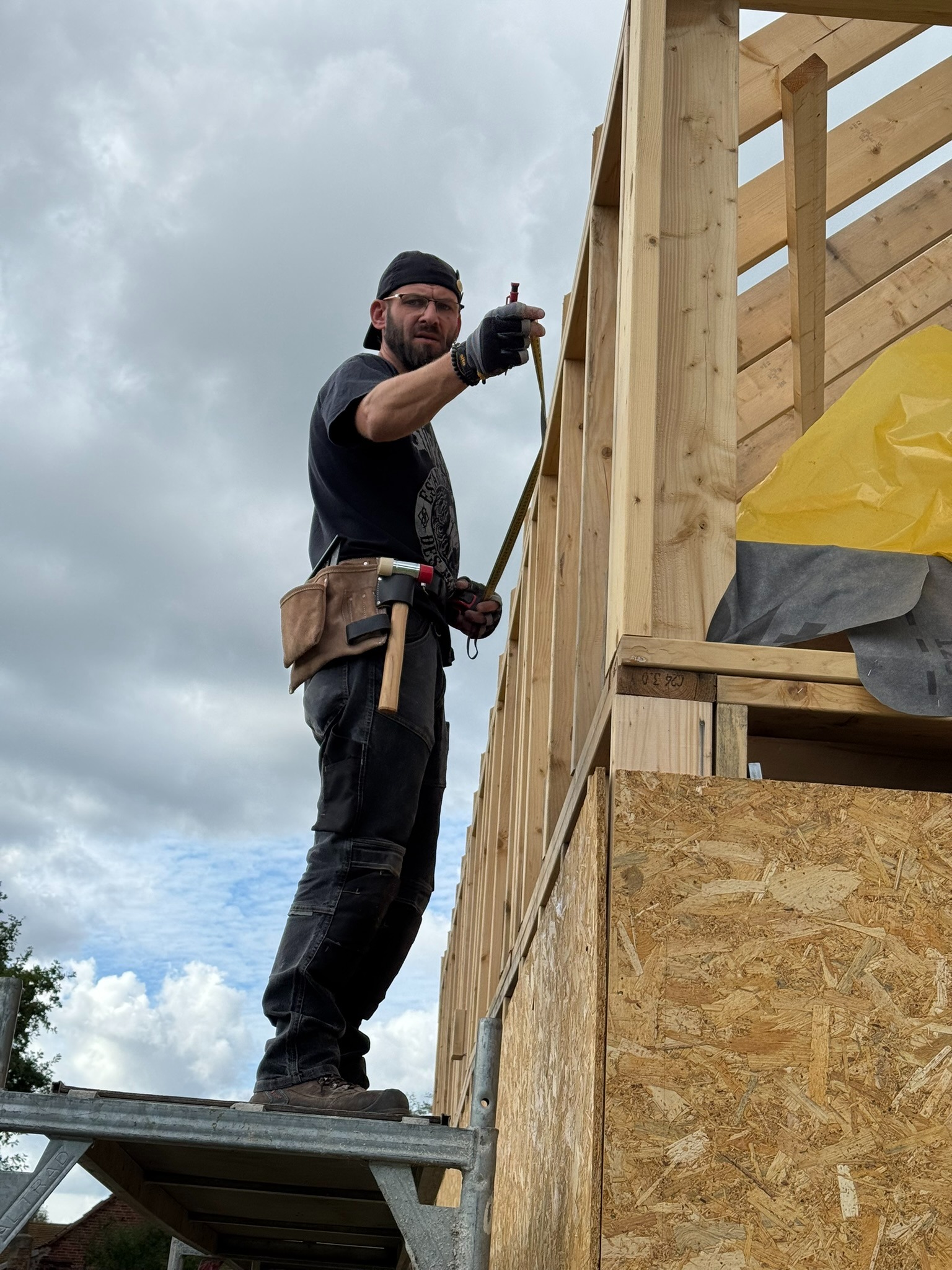 Monter w czapce, mierzy konstrukcję domu szkieletowego. Widoczne drewniane elementy, płyty OSB i narzędzia. Ujęcie z dołu, na tle pochmurnego nieba.