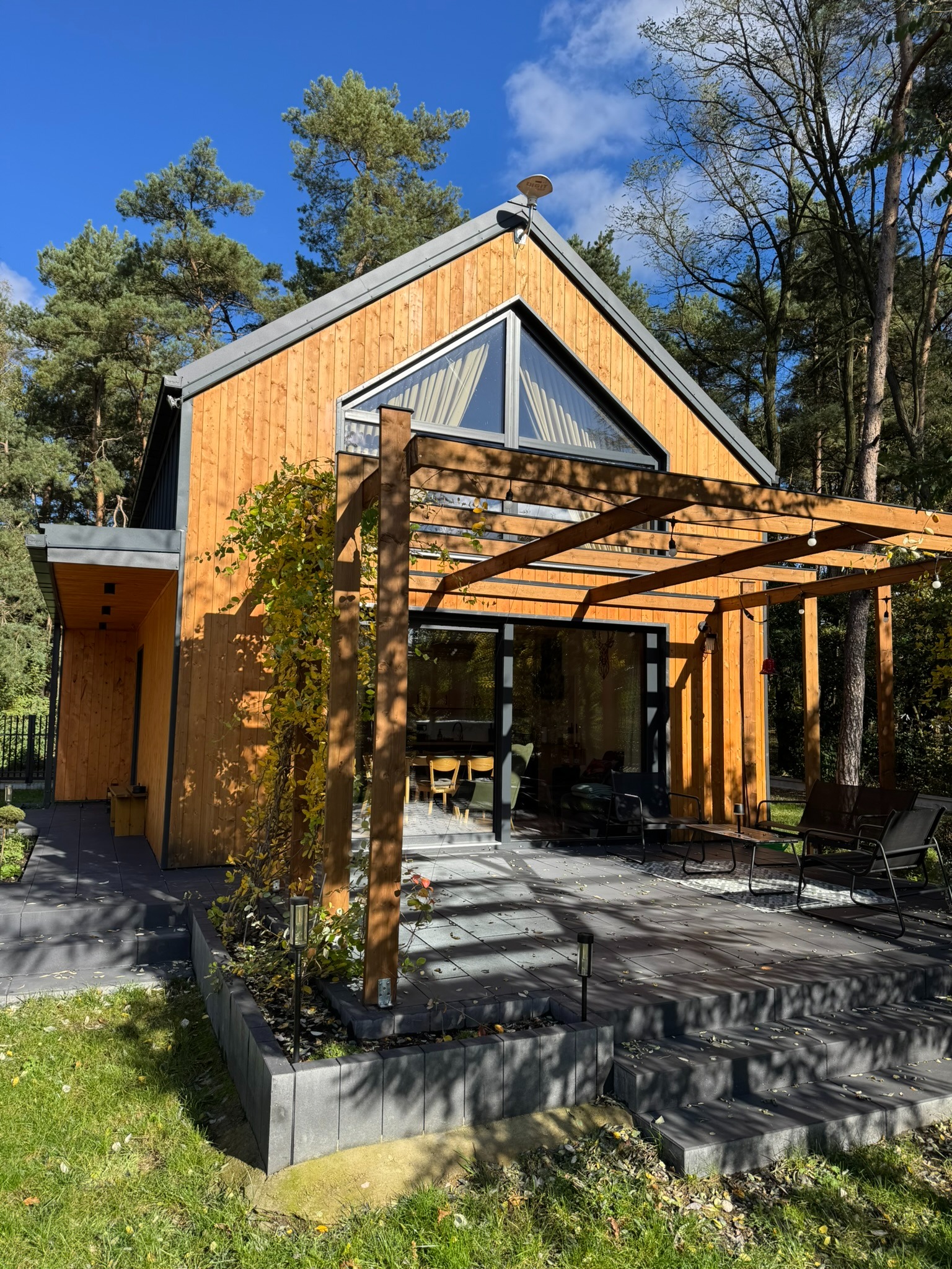 Nowoczesny dom szkieletowy z drewnianą elewacją i pergolą, otoczony zielenią. Duże okno trójkątne w szczycie. Taras z meblami ogrodowymi.