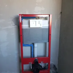 Stelaż podtynkowy WC z czerwoną ramą, niebieskimi rurami i czarnym odpływem, zamontowany na surowej ścianie w trakcie prac instalacyjnych.