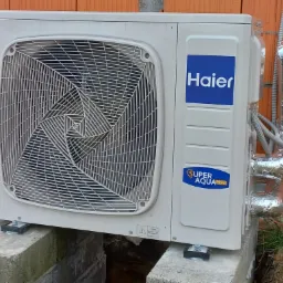Zewnętrzna jednostka klimatyzacji Haier zamontowana na betonowych blokach przy ceglanym murze, z widocznymi izolowanymi rurami.