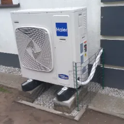 Biała pompa ciepła Haier zamontowana na betonowych podstawach przy ścianie budynku z szarą podmurówką i kamiennym podsypem. Widoczna metalowa siatka ochronna.