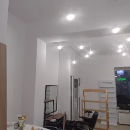 Wnętrze salonu fryzjerskiego w trakcie prac wykończeniowych; widoczne białe ściany, sufit z podwieszonym oświetleniem, stanowisko fryzjerskie z narzędziami na blacie oraz regały w tle.