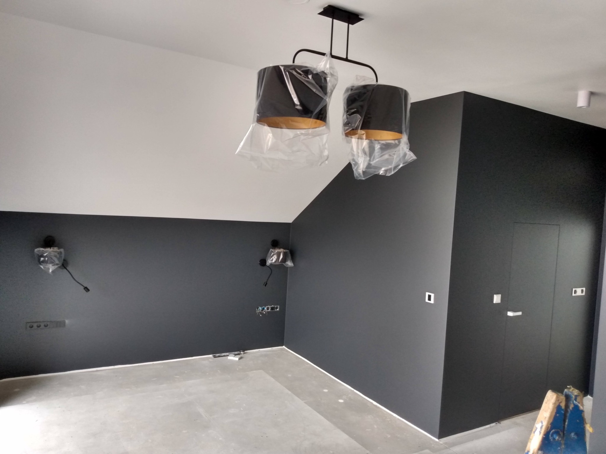 Pokój po remoncie z ciemnymi ścianami, białym sufitem, nową lampą w folii ochronnej i widocznymi puszkami elektrycznymi.