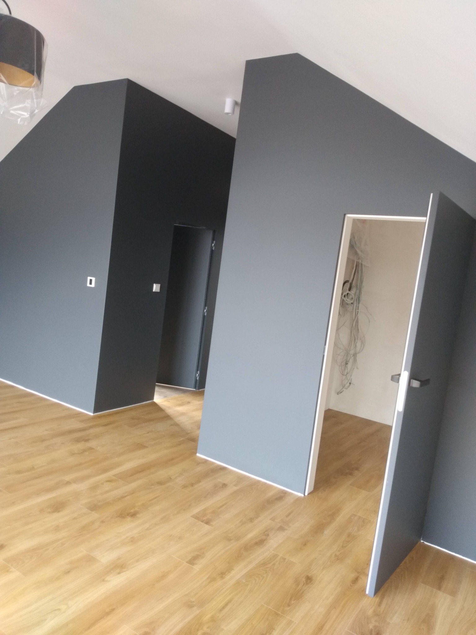 Pomieszczenie po remoncie: szare ściany, drewnopodobna podłoga, otwarte drzwi z widoczną instalacją elektryczną w środku.