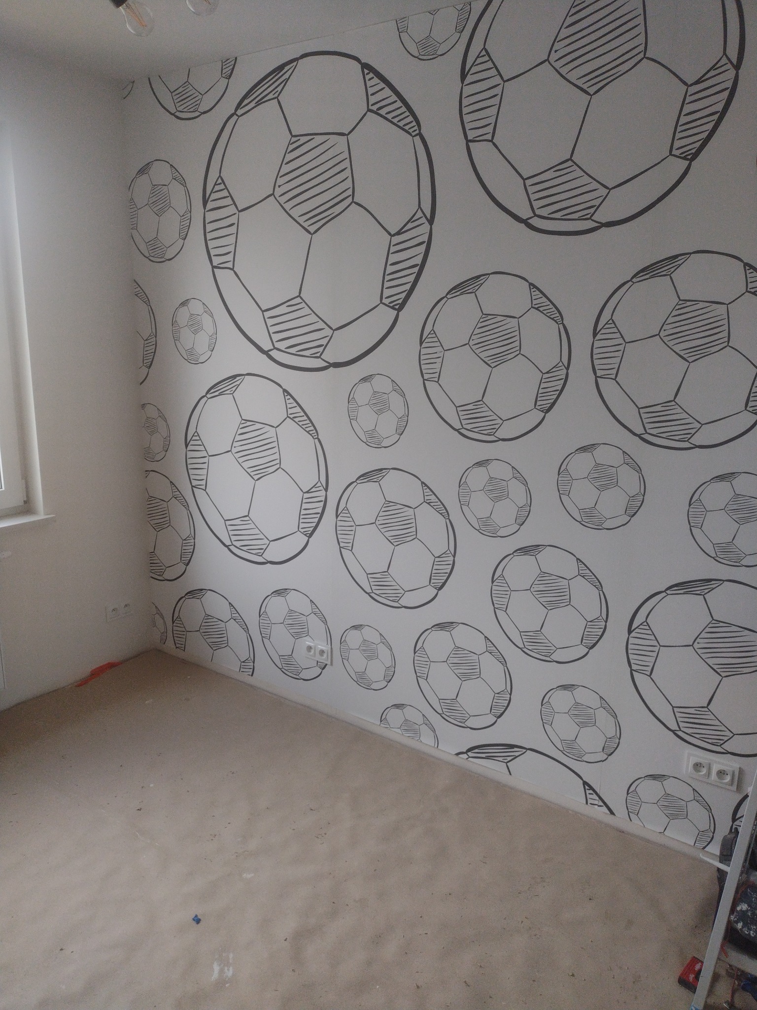 Ściana pokoju dziecięcego z ręcznie malowanym wzorem piłek nożnych w czerni na białym tle, widoczne gniazdka elektryczne i fragment podłogi przed wykończeniem.