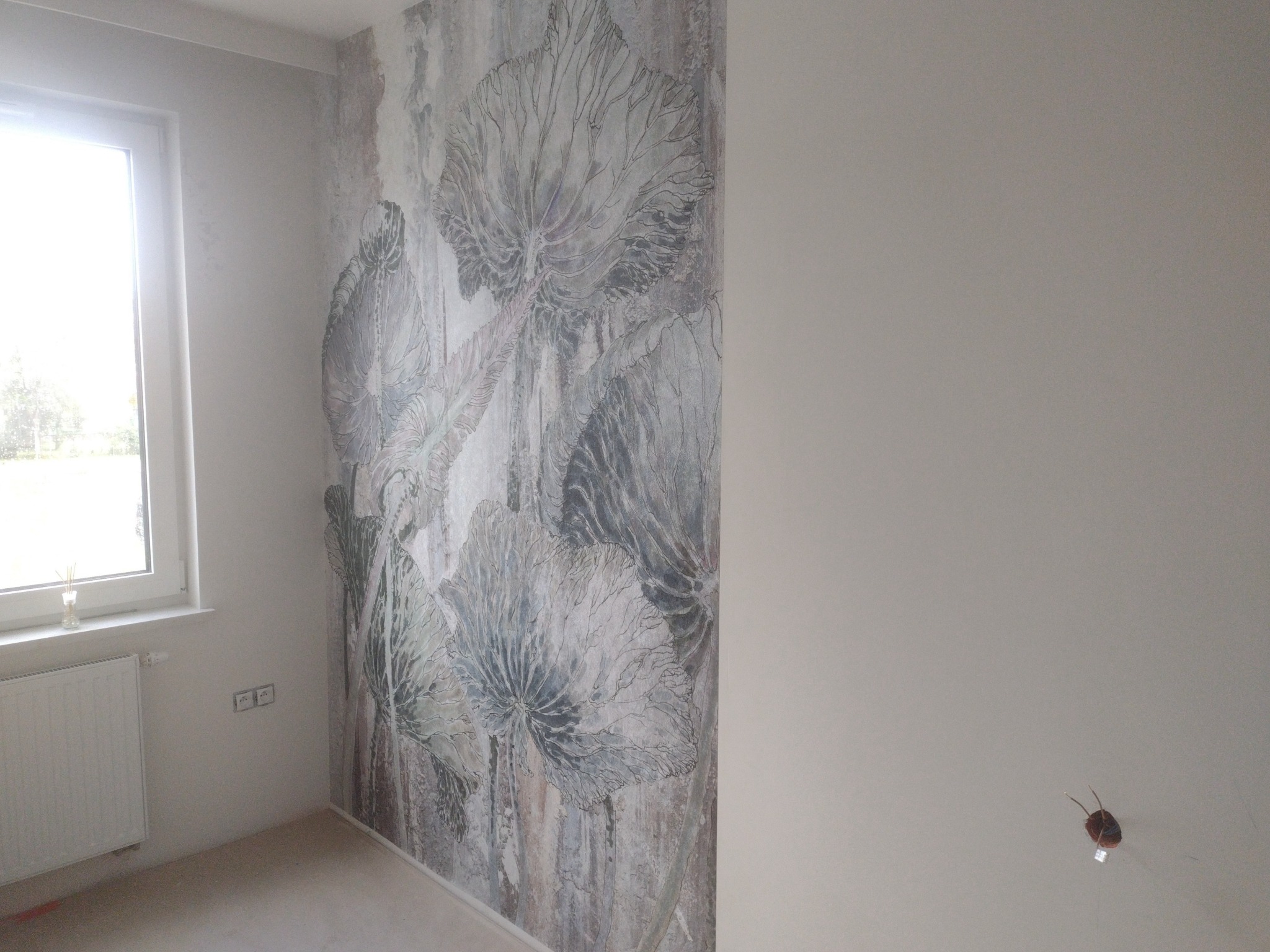 Ściana w pokoju z nową tapetą w motywy roślinne, obok ściana pomalowana na jasny kolor, widoczne gniazdka elektryczne i grzejnik pod oknem.