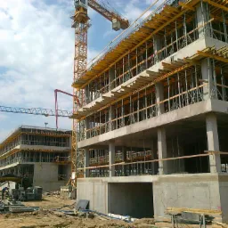 Wielopiętrowy budynek w surowym stanie, z widocznym szalunkiem i podporami, na tle placu budowy z dwoma żurawiami wieżowymi i pracownikami w oddali.