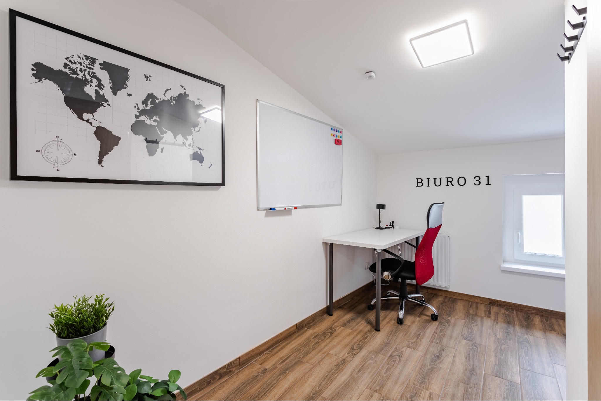 Jasne wnętrze biura z biurkiem, krzesłem z czerwonym oparciem, tablicą suchościeralną, mapą świata i roślinami doniczkowymi, oświetlone nowoczesną lampą sufitową.