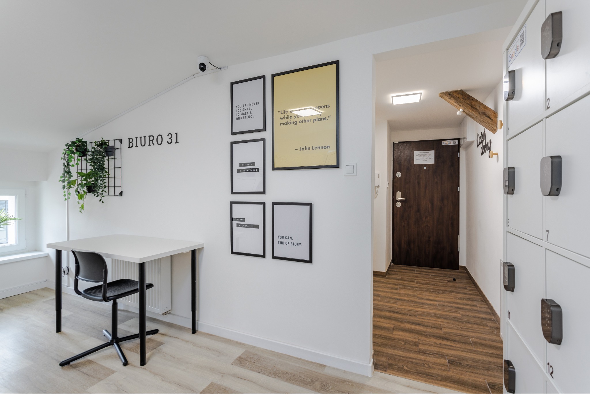 Jasne wnętrze biurowe: białe ściany z dekoracyjnymi plakatami motywacyjnymi, biurko z krzesłem, szafki depozytowe z numerami oraz rośliny doniczkowe na ścianie.