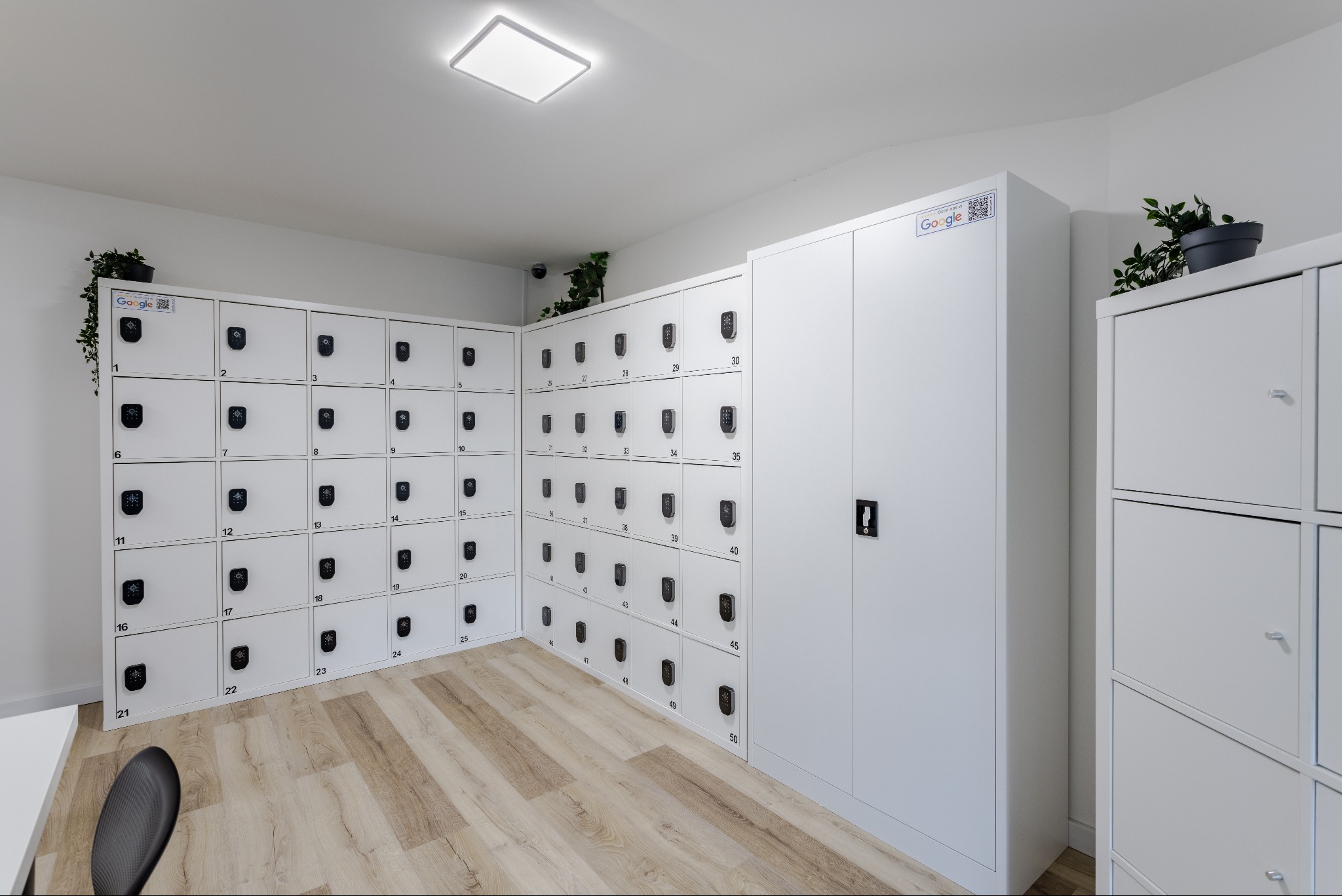 Jasne pomieszczenie z rzędem białych szafek z elektronicznymi zamkami, oznaczonych numerami, z naklejkami Google, na podłodze panele imitujące drewno.
