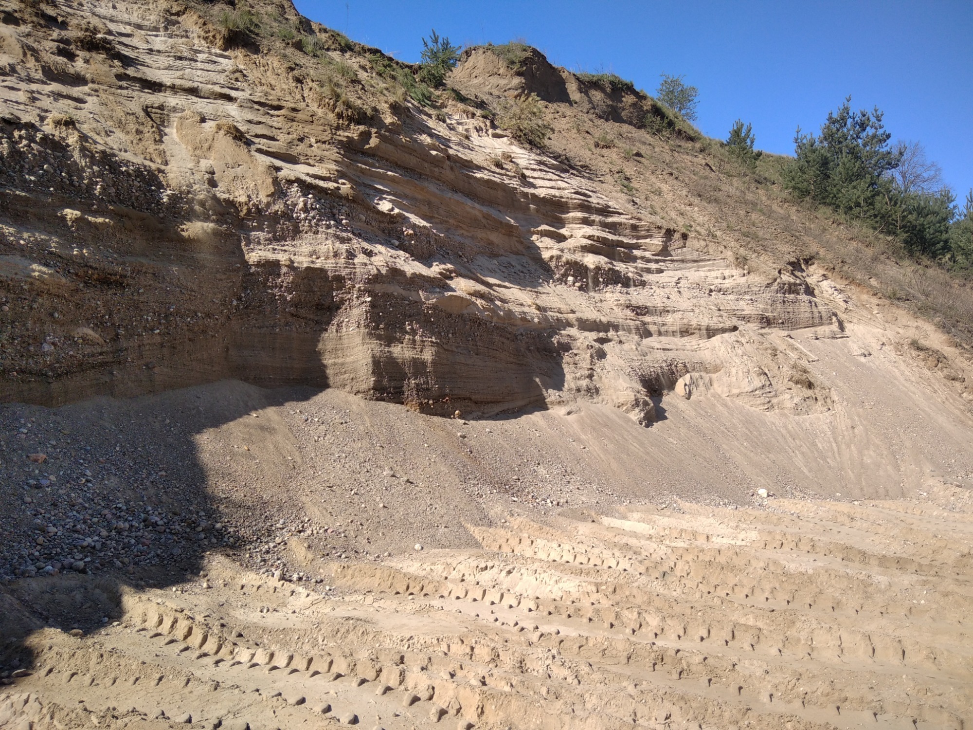 Skarpa ziemna z widocznymi warstwami gruntu i śladami opon ciężkiego sprzętu na dole, sugerująca prowadzone prace ziemne.