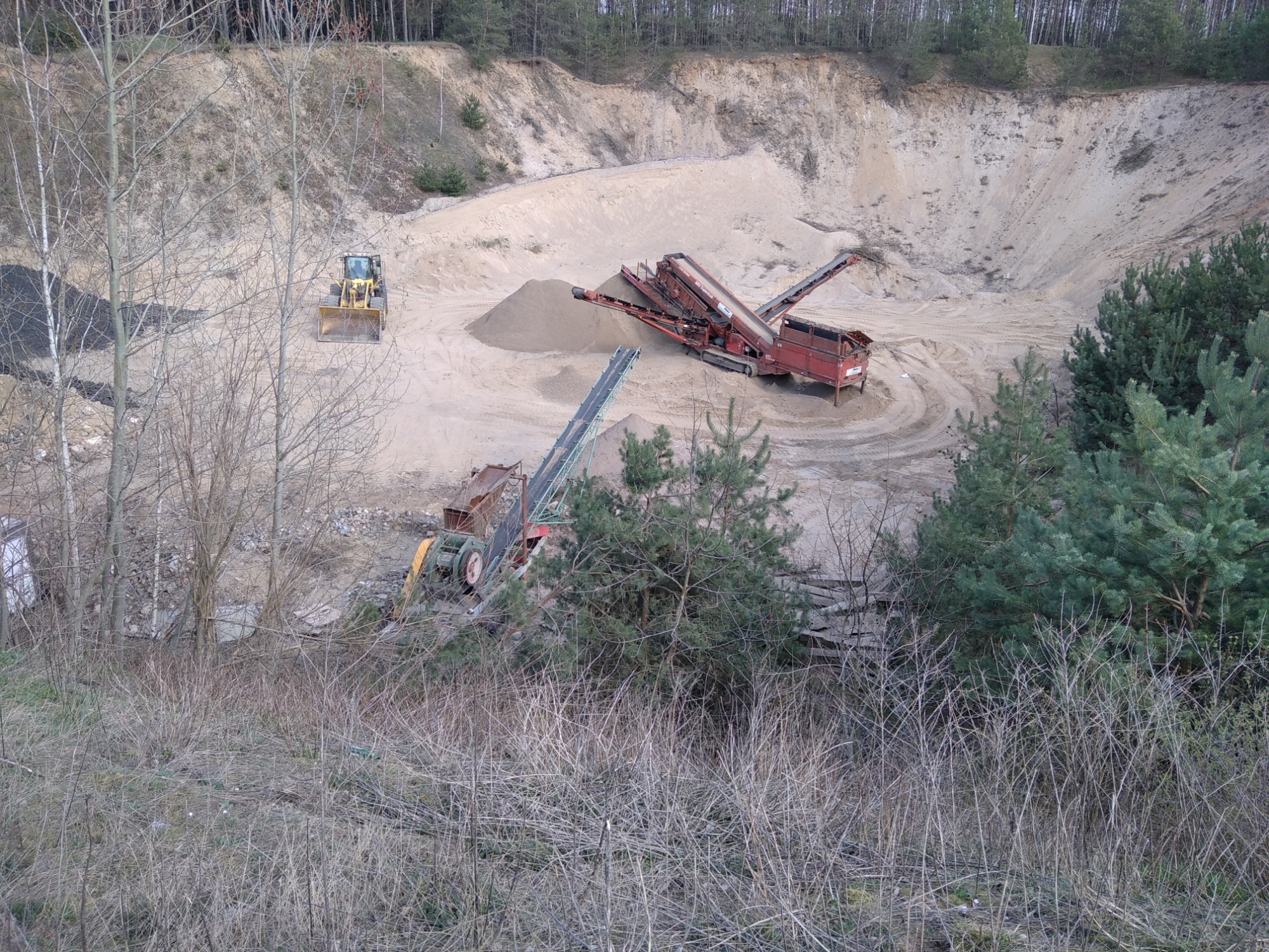 Widok z góry na wyrobisko piasku z pracującym separatorem kruszywa i ładowarką kołową w tle, otoczone brzegiem wyrobiska i drzewami.