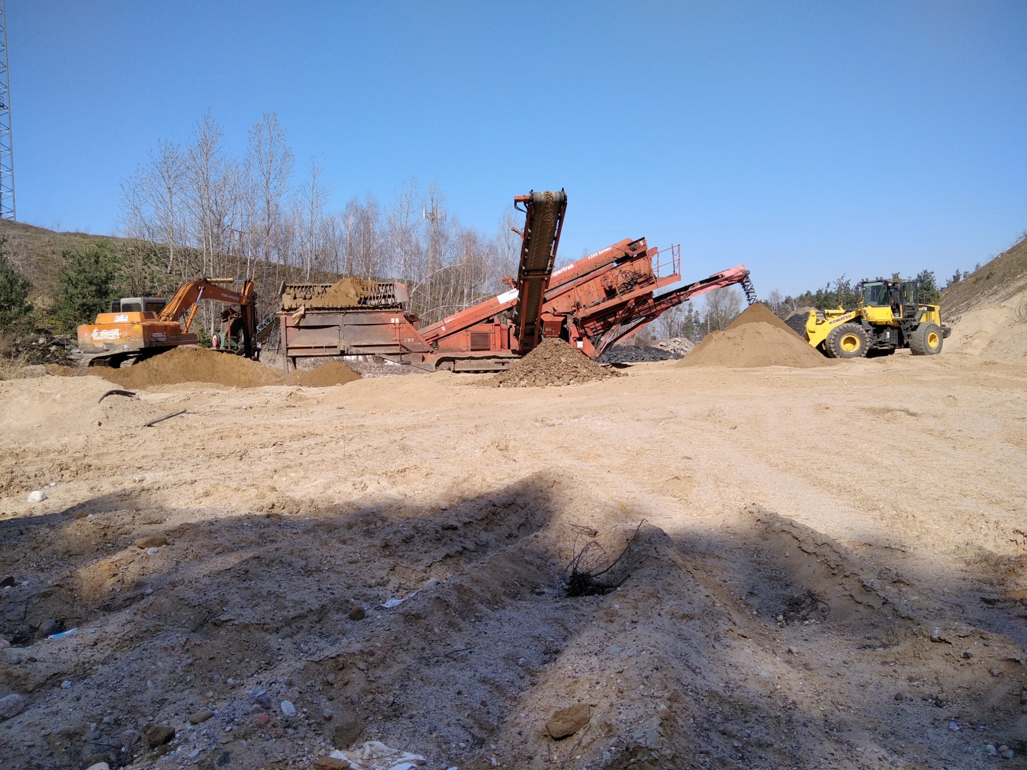 Żółta ładowarka kołowa i pomarańczowa koparka pracują przy przesiewaczu do ziemi na placu budowy w słoneczny dzień. Widoczne pryzmy piasku i ziemi.