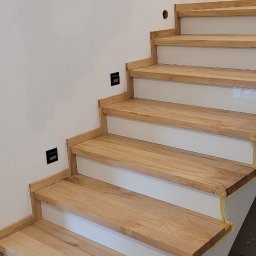 Stanisław Strzęp - Montaż drewnianych schodów z jasnego drewna na białej konstrukcji, zabezpieczonych taśmą malarską, widoczne gniazdka elektryczne na ścianie.
