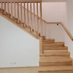 Jasne drewniane schody z balustradą w minimalistycznym wnętrzu, widoczne gniazdka elektryczne przy podłodze i odbicie schodów w szklanych drzwiach.