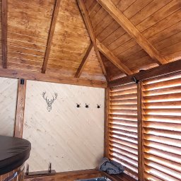 Stanisław Strzęp - Wnętrze drewnianej altany z ażurowymi ścianami, widoczne trzy wieszaki na jasnej ścianie z motywem głowy jelenia, drewniana ławka i fragment beczki.