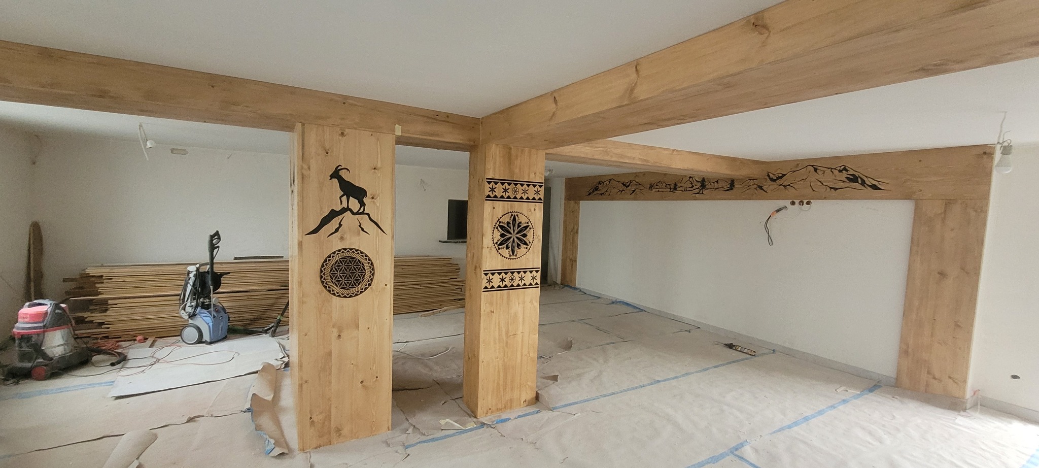 Surowe drewniane słupy i belki w pokoju z motywami góralskimi, zdobione czarnymi sylwetkami kozic i wzorami kwiatowymi, podłoga zabezpieczona folią malarską, widoczne narzędzia i materiały...