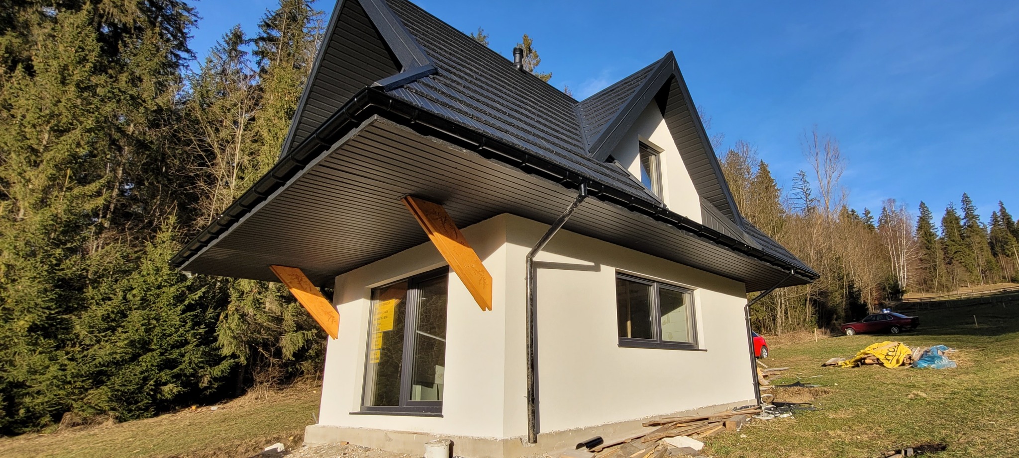 Nowoczesny dom z białą elewacją, czarnym dachem i drewnianymi elementami wspierającymi okap, na tle lasu w słoneczny dzień.