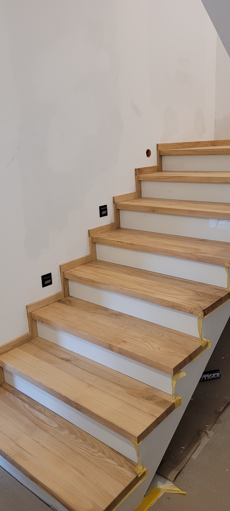 Montaż drewnianych schodów z jasnego drewna na białej konstrukcji, zabezpieczonych taśmą malarską, widoczne gniazdka elektryczne na ścianie.
