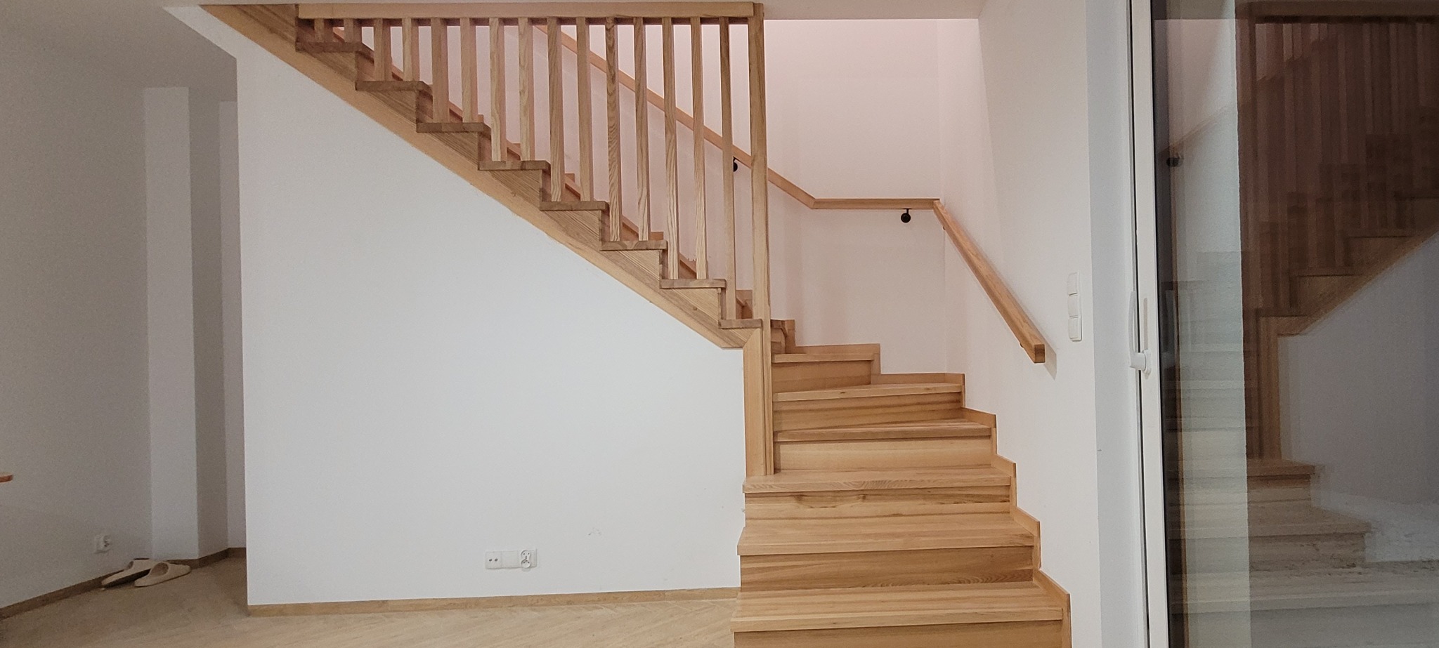 Jasne drewniane schody z balustradą w minimalistycznym wnętrzu, widoczne gniazdka elektryczne przy podłodze i odbicie schodów w szklanych drzwiach.