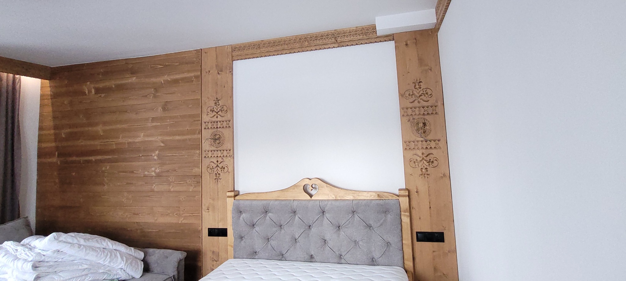 Pokój z drewnianą boazerią i ramą łóżka z motywami góralskimi, szara tapicerowana rama łóżka z wycięciem w kształcie serca, biała ściana