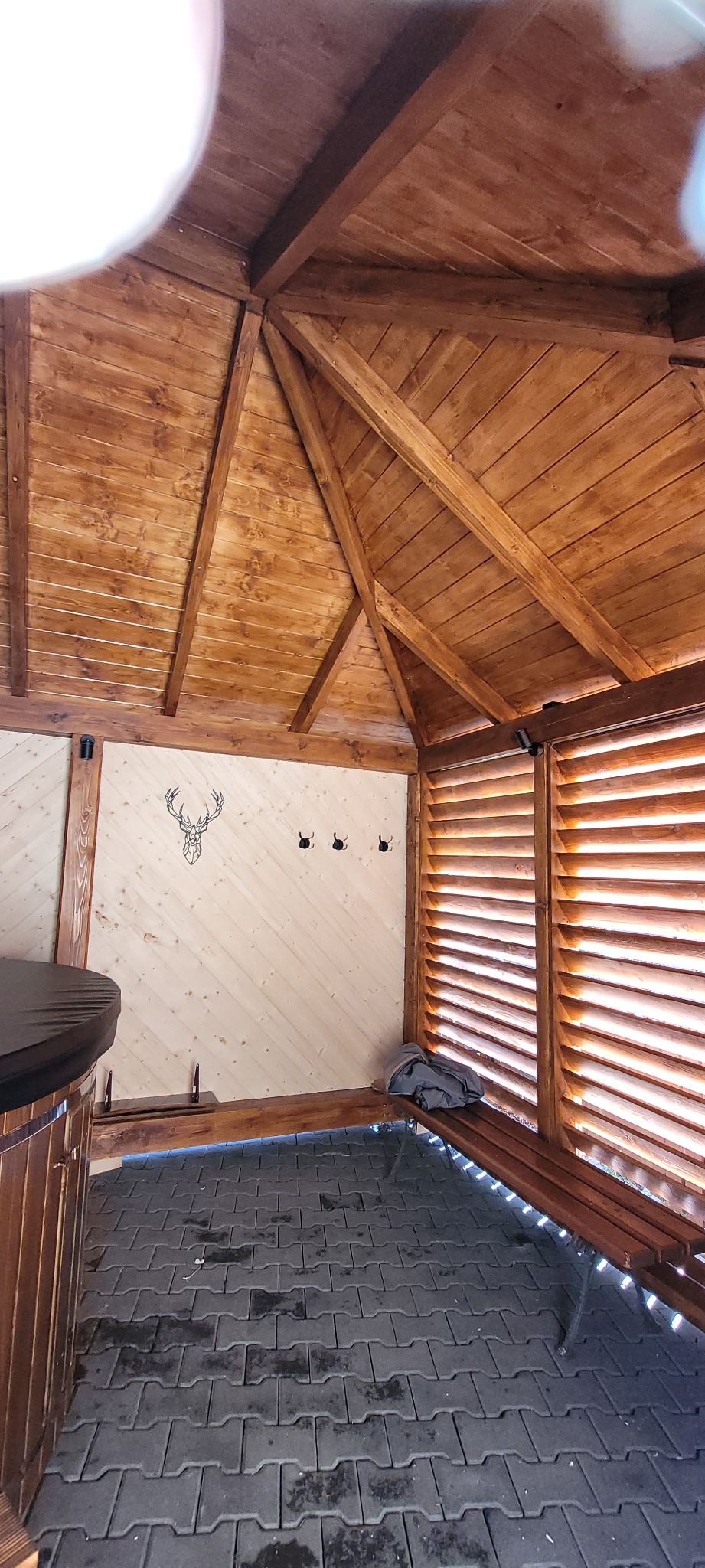 Wnętrze drewnianej altany z ażurowymi ścianami, widoczne trzy wieszaki na jasnej ścianie z motywem głowy jelenia, drewniana ławka i fragment beczki.