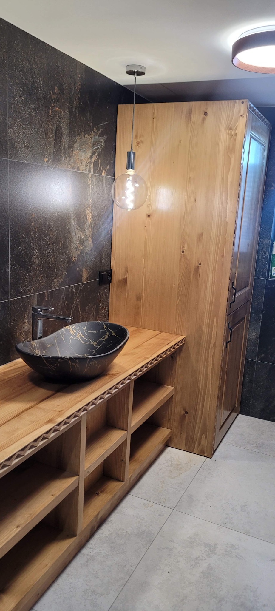 Nowoczesna łazienka z drewnianą szafką pod umywalkę i czarną umywalką z marmurkowym wzorem. Na ścianie ciemne płytki imitujące kamień. Wisząca lampa z żarówką Edisona.