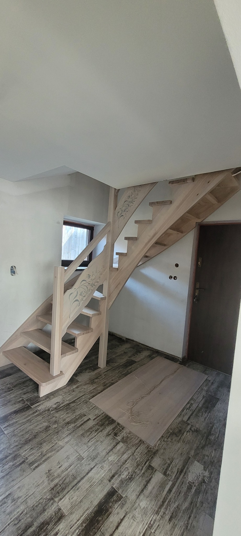 Jasne, drewniane schody z dekoracyjnymi wycięciami, prowadzące na górę w nowym wnętrzu. Widoczne wykończenie podłogi imitujące drewno i fragment drzwi.
