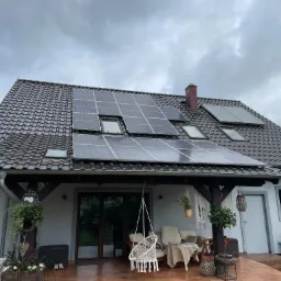 Dom z panelami słonecznymi na dachu z dachówki ceramicznej, widoczne okna dachowe, taras z meblami ogrodowymi i wiszącym fotelem, pochmurne niebo.