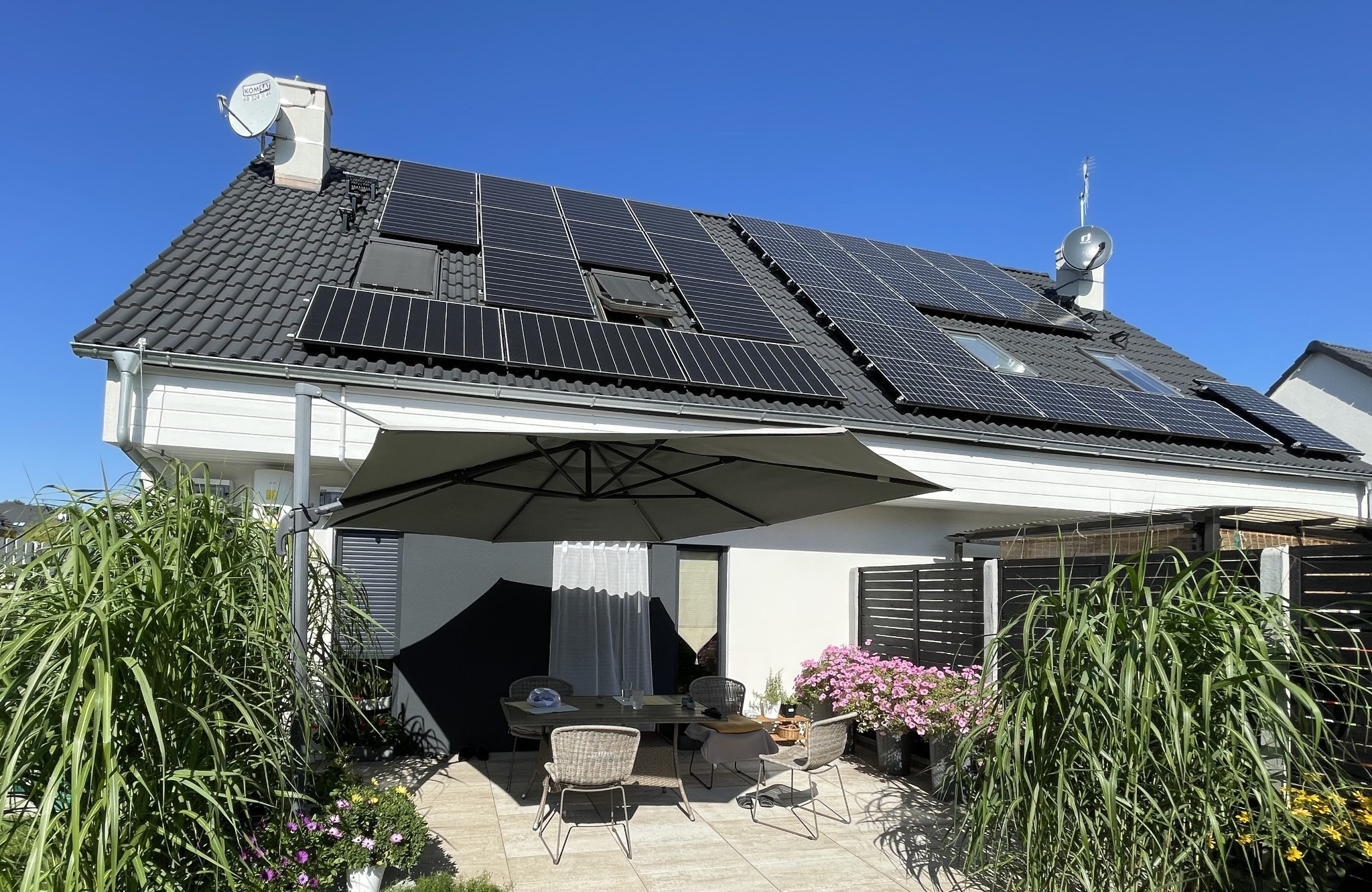 Dom jednorodzinny z panelami słonecznymi na dachu, widoczne z poziomu tarasu z parasolem i roślinnością.