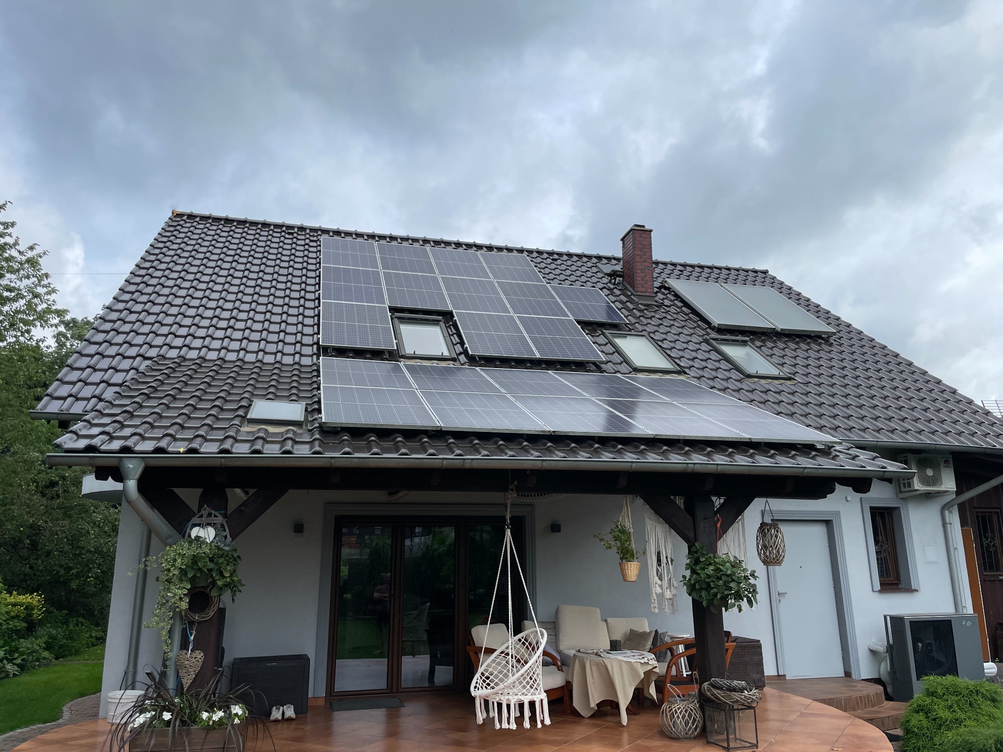 Dom z panelami słonecznymi na dachu z dachówki ceramicznej, widoczne okna dachowe, taras z meblami ogrodowymi i wiszącym fotelem, pochmurne niebo.