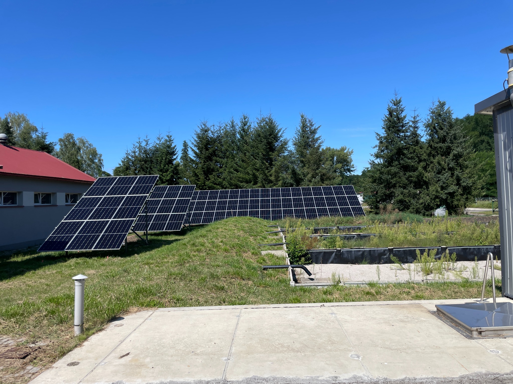 Instalacja fotowoltaiczna na trawiastym terenie z systemem odwadniającym, widoczne panele słoneczne i elementy konstrukcyjne, w tle budynek z czerwonym dachem i drzewa iglaste.