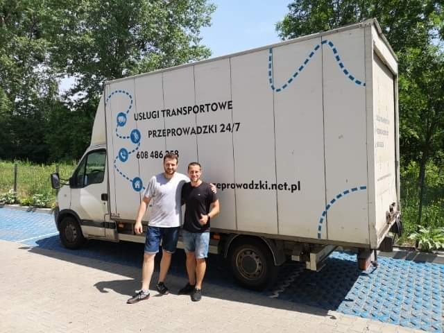 Dwóch mężczyzn pozuje przed białym samochodem dostawczym z napisem 'Usługi Transportowe Przeprowadzki 24/7' i numerem telefonu, zaparkowanym na niebieskiej kostce brukowej w słoneczny dzień.