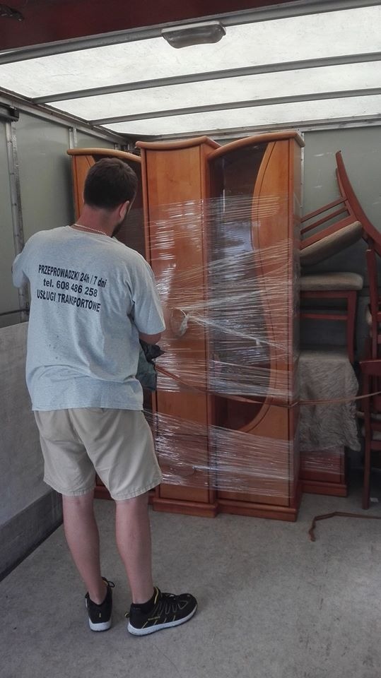 Mężczyzna w koszulce z nadrukiem firmy przeprowadzkowej zabezpiecza folią stretch drewniany, przeszklony kredens wewnątrz przestrzeni ładunkowej, obok ustawione krzesła.