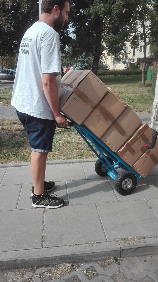 Mężczyzna w krótkich spodenkach transportuje kartony na niebieskim wózku dwukołowym po chodniku.