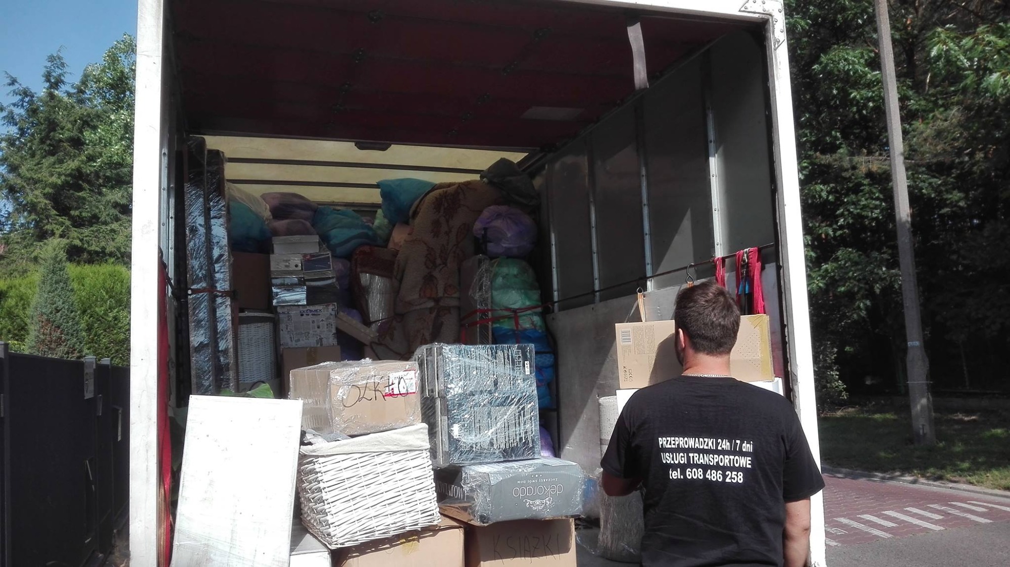 Wnętrze samochodu dostawczego wypełnione po brzegi spakowanymi kartonami, meblami i kocami, widok z tyłu na mężczyznę w czarnej koszulce z nadrukowanym numerem telefonu firmy transportowej.