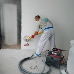 Pracownik w kombinezonie i masce szlifuje ścianę szlifierką z odkurzaczem, przygotowując ją do dalszych prac wykończeniowych; widoczne zabezpieczone folią drzwi.